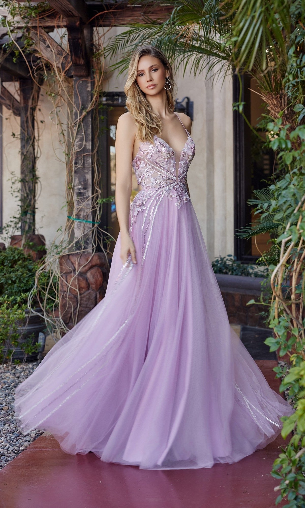 A woman wears the Nox Anabel G1795 long pink prom dress with an a-line tulle skirt and floral embellishments, standing outdoors by a building and greenery.